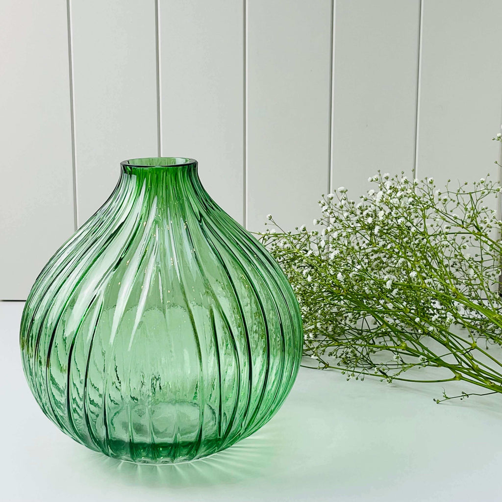 A glass fluted vase with a grooved pattern. The fluted vase is green in colour and is placed in front of a bunch of gyp
